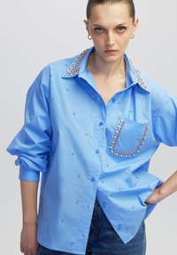Blauw oversized button-up shirt met versierde strassdetails op de kraag en zak, met een gladde textuur en ontspannen mouwen.