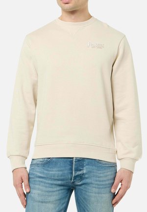 Mann trägt beige Sweatshirt mit „Boxeur Des Rues“ Logo auf der linken Brust und blaue Jeans, steht vor weißem Hintergrund.