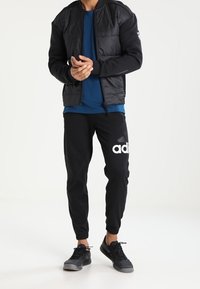 Veste bomber matelassée noire, chemise bleue, joggers noirs avec logo blanc, et baskets noires. Texture lisse, design ajusté avec poignets élastiques.