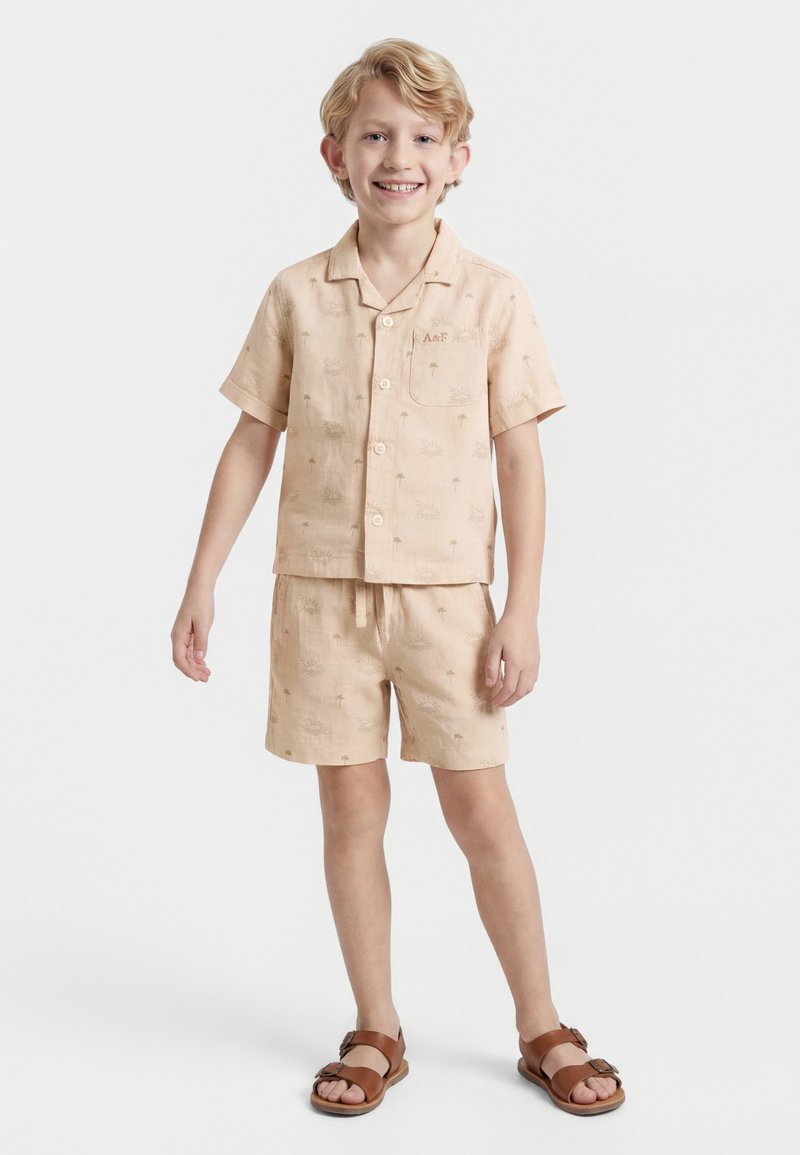 Χαμογελαστός νεαρός αγόρι με ξανθά μαλλιά, φοράει μπεζ κοντομάνικο πουκάμισο με κουμπιά, κοντά παντελόνια με στάμπα φοίνικα και καφέ σανδάλια, όρθιος.