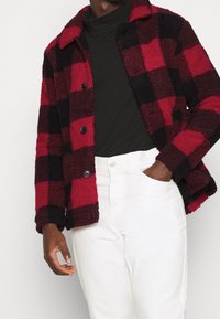Red and black checkered fleece jacket with a button-up front, spread collar, and textured fabric. Worn over a black turtleneck and white pants.