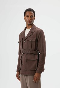 Braune Jacke aus Wollmischung mit strukturiertem Muster, doppelreihiger Vorderseite, vier aufgesetzten Taschen und einem Taillengürtel. Mit Reverskragen.