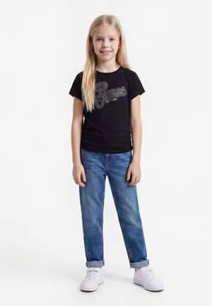 Ung pige med langt blondt hår iført en sort Guess t-shirt, blå opkørte jeans og hvide sneakers, står og smiler fremad.