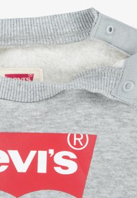 Grå hettegenser med ribbet krage, trykknapper og stor rød Levi's-logo. Myk, fleece-lignende innvendig fôr.