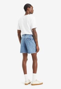 Shorts din denim de culoare albastru deschis, cu o croială lejeră, purtate cu un tricou alb. Caracteristici includ două buzunare spate și cusături vizibile.