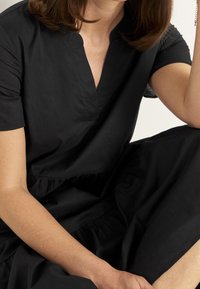 Schwarzes Kleid mit kurzen Ärmeln, V-Ausschnitt und abgestufter, geraffter Saumkante. Der Stoff wirkt glatt mit einer leichten Textur.