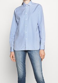 Blue and white striped long-sleeve shirt with a button-down collar, tailored fit, and curved hem, paired with light blue jeans.