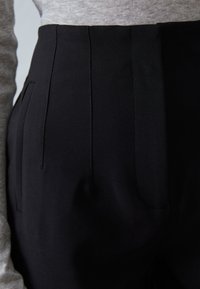 Pantalon noir sur mesure avec des plis verticaux, des poches latérales et une texture lisse, présenté avec un haut gris clair.