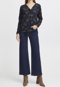 Kvinde iført en marineblå blomstret bluse med lange ærmer, mørke bukser med vide ben og brune støvler med hæl, stående med den ene hånd i lommen.