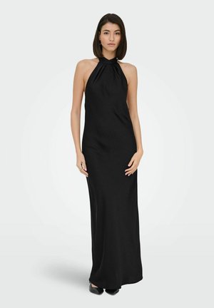 Femme aux cheveux raides mi-longs, portant une robe longue noire sans manches à col halter et des chaussures noires pointues, debout.