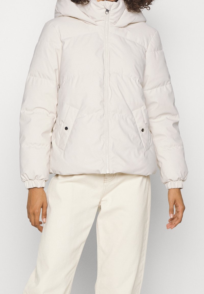 Osoba w kremowej, pikowanej kurtce z elastycznymi mankietami i zapięciem na zamek z przodu, w połączeniu z jasnobeżowymi spodniami na jednolitym tle.