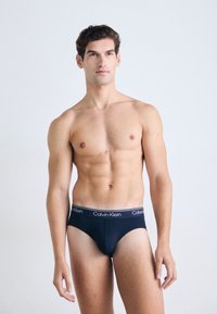 Fit männliches Modell steht in schwarzen Calvin Klein Unterhosen, leicht zur Seite blickend, vor einem schlichten hellen Hintergrund.