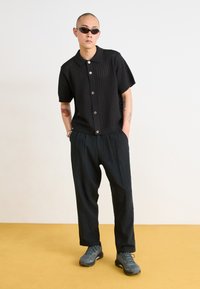 Black short-sleeve knit shirt with button front, textured fabric, and loose-fitting black trousers paired with gray hiking shoes.