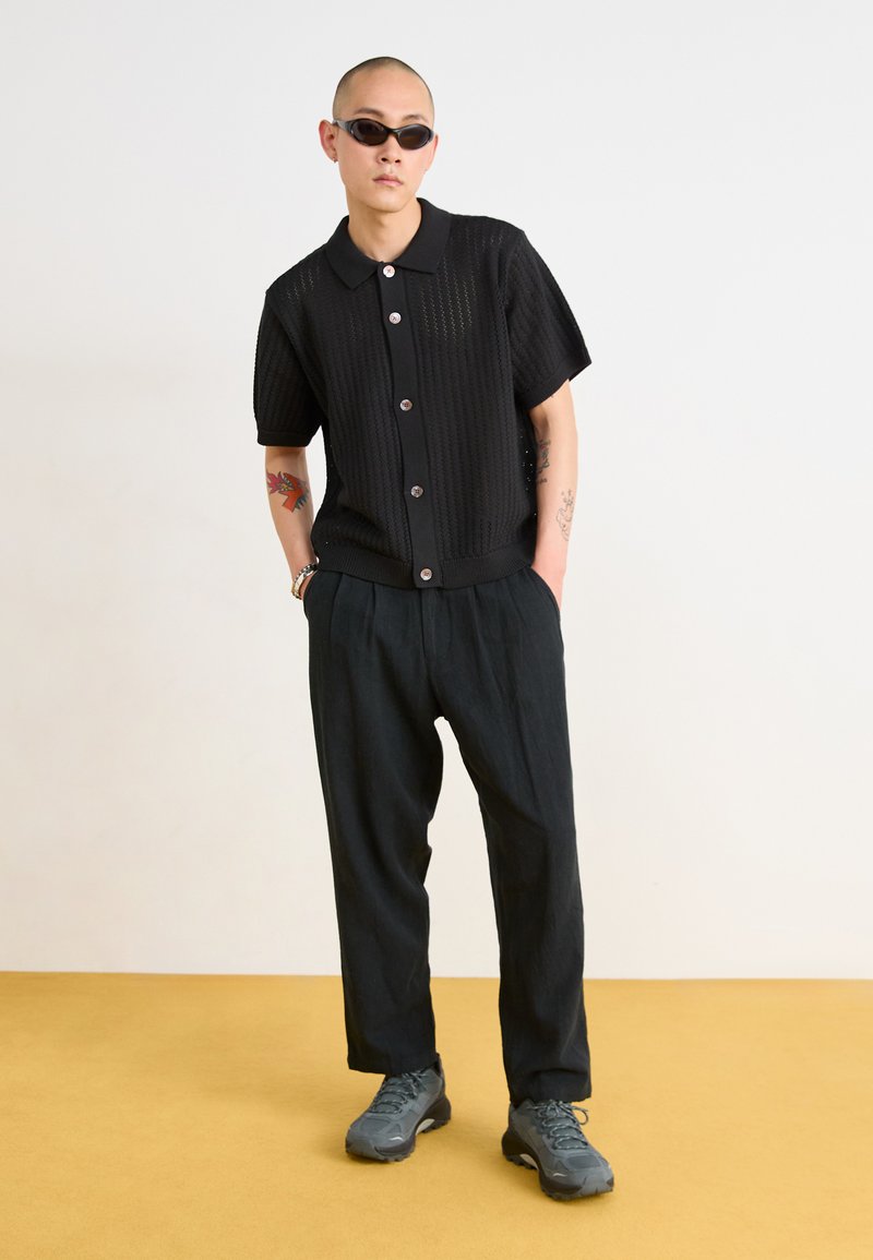 Black short-sleeve knit shirt with button front, textured fabric, and loose-fitting black trousers paired with gray hiking shoes.