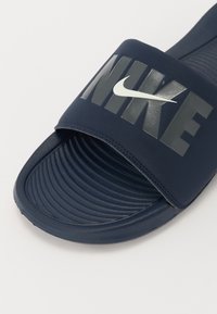 Sandale de plajă bleumarin cu o bandă largă, având o marcaj mare "NIKE" de culoare gri, logo al aripii alb și o talpă texturată pentru confort.