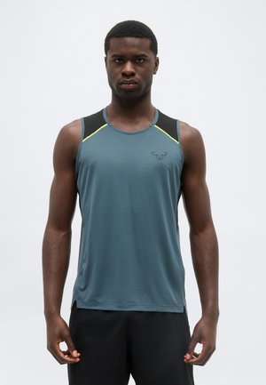 Athletischer Mann trägt ein ärmelloses blau-graues Sportshirt mit schwarzen und gelben Akzenten, steht vor einem schlichten hellen Hintergrund.