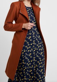 Abrigo largo y ajustado de color óxido, hecho de una tela texturizada, con un cuello de muesca y un cierre metálico. Combinado con un vestido floral azul marino.