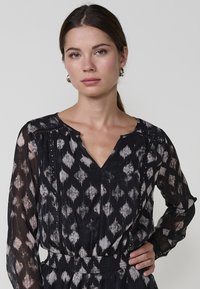 Schwarzes und cremefarbenes gemustertes Kleid mit V-Ausschnitt, langen durchsichtigen Ärmeln, elastischem Taillenbund und dekorativen Perlen entlang des Ausschnitts.