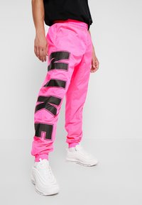 Person wearing bright pink Nike joggers with large black Nike logo on leg and white Nike sneakers against plain background.