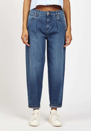 Woman wearing high-waisted, relaxed fit blue jeans with rolled cuffs and white sneakers, standing against a plain background.