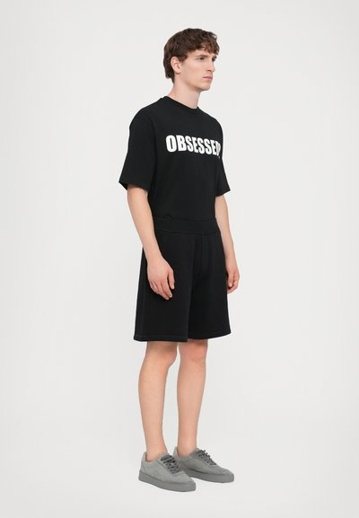 T-shirt en coton noir avec "OBSESSED" en texte blanc, associé à un short noir et des baskets en suède gris. Tenue athlétique simple et décontractée.