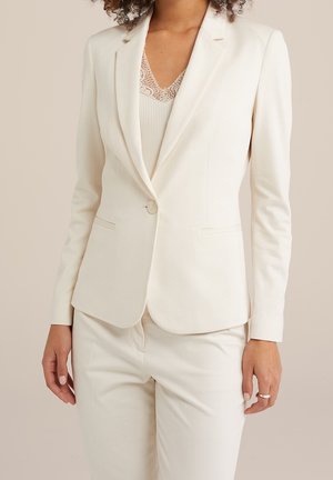 Kvinde iført en off-white skræddersyet blazer med en enkelt knap og matchende bukser, kombineret med en ribbet camisole med blonder.