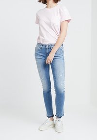 Camiseta de algodón rosa claro con mangas cortas y un bolsillo, combinada con unos vaqueros skinny desgastados de color azul claro y zapatillas blancas.