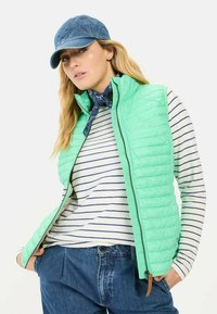Hellgrüne Pufferweste mit horizontalem Steppmuster, mit Reißverschluss; getragen über einem gestreiften Langarmshirt in Weiß und Marineblau; kombiniert mit blauen Jeans.