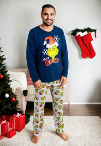 Maglietta a maniche lunghe blu navy con grafica del Grinch e testo "TEAM GRINCH"; pantaloni da pigiama grigi con motivo colorato del Grinch; a piedi nudi.