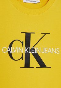 Tejido amarillo con un gran logo negro "cK" y el logo blanco "CALVIN KLEIN JEANS" impreso en el centro.
