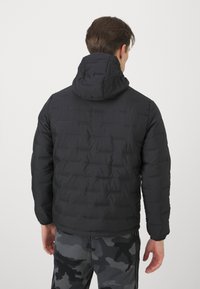 Giacca nera imbottita con cappuccio, caratterizzata da un design testurizzato e trapuntato e polsini elastici. Abbinata a pantaloni grigi in camouflage.