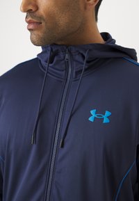 Marineblå zip-opp-jakke laget av elastisk stoff, med høy krage, justerbare snorer og en kontrasterende blå logo på brystet.