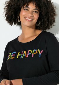 Donna sorridente con capelli ricci, che indossa orecchini a cerchio e una maglietta nera con la scritta colorata "BE HAPPY" sul petto.
