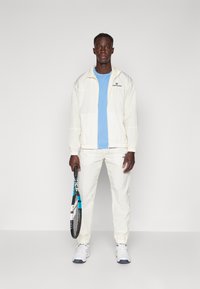 Hvid sportsjakke med en blå t-shirt og matchende bukser, stoffet ser letvægtigt ud, slankt design, parret med en tennisketsjer.