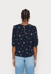 Marineblauwe bloemenblouse met pofmouwen, voorzien van kleurrijk borduurwerk en een open rugdetail met een sleutelgat. Gecombineerd met lichtblauwe spijkerbroek.