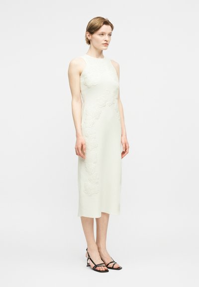 Γυναίκα που φοράει ένα λευκό μίντι φόρεμα χωρίς μανίκια με κεντήματα λουλουδιών και μαύρα ψηλοτάκουνα σανδάλια με λουράκια, στέκεται μπροστά σε ένα απλό φόντο.