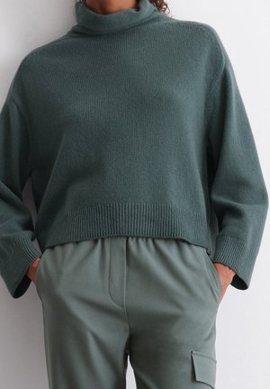 Personne portant un pull en maille vert foncé à col montant et un pantalon vert assorti avec des poches latérales, les mains dans les poches.