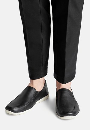 Schwarze Leder-Slipper mit strukturiertem Finish und weißen Gummi-Sohlen, kombiniert mit schwarzen, knöchelfreien Hosen.