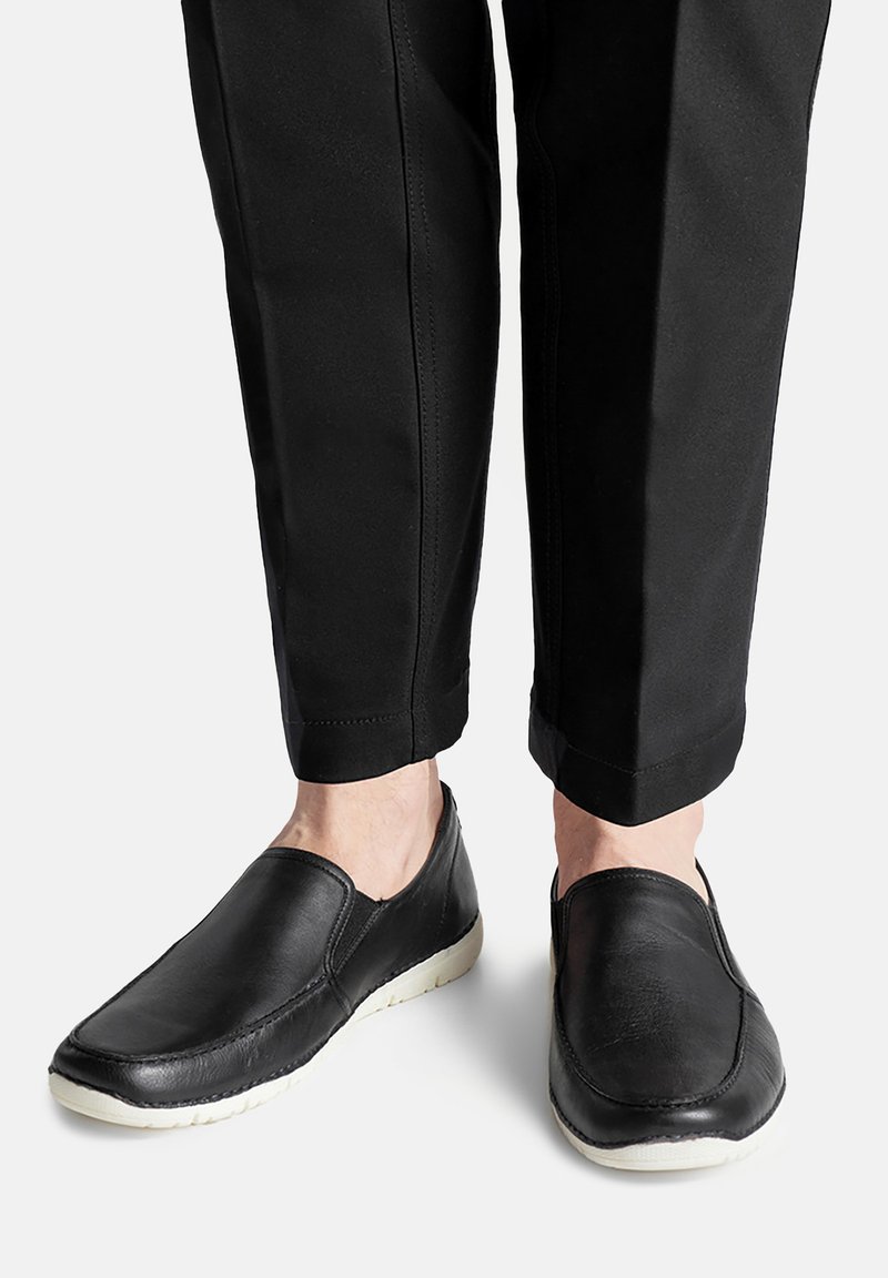 Czarne skórzane buty wsuwane z teksturowanym wykończeniem i białymi gumowymi podeszwami, zestawione z przyciętymi czarnymi spodniami.