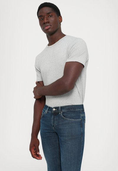 Giovane uomo che indossa una maglietta a maniche corte grigia chiaro e jeans blu scuro, in piedi contro uno sfondo bianco uniforme con le braccia incrociate.