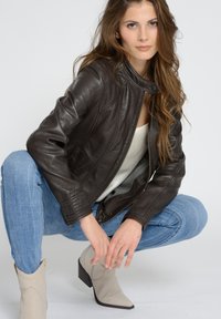 Brun läderjacka med ribbade detaljer, dragkedja framtill och ståkrage, i kombination med ljusblå jeans och beiga ankelboots.