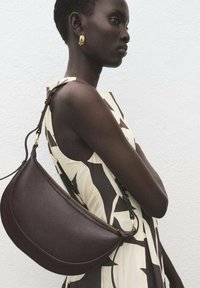 Bolso de cuero marrón en forma de media luna con una superficie texturizada y cierre de cremallera, llevado en bandolera sobre un vestido blanco con estampado.