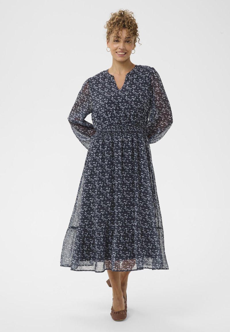 Femeie zâmbitoare cu păr creț, îmbrăcată într-o rochie midi cu flori, de culoare bleu marin, cu mâneci lungi din material transparent, stând cu mâinile în spate.