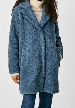 Personne portant un manteau en polaire texturé bleu mi-long par-dessus un pull à col roulé crème et un jean bleu, les mains dans les poches du manteau.