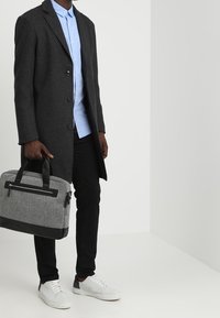 Grå rock, ljusblå skjorta, svarta byxor, vita sneakers. Håller en grå och svart texturerad väska med dragkedjor och handtag. Enkel, modern design.