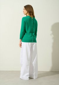 Zielony sweter z dzianiny o okrągłym dekolcie, z guzikiem z tyłu, w zestawieniu z białymi spodniami typu wide-leg.