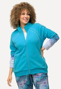 Sudadera turquesa con cremallera y costuras blancas, de manga larga con puños estampados; combinada con pantalones multicolores de patrón mármol.