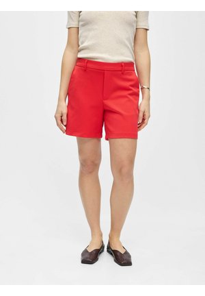 Femme portant un short rouge vif sur mesure, un haut beige à manches courtes, des ballerines noires et un bracelet en argent, debout devant un fond uni.