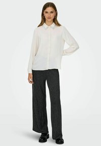 Blouse blanche à manches longues avec un col et des boutons, associée à un pantalon large gris foncé texturé. Chaussures plates avec des clous.