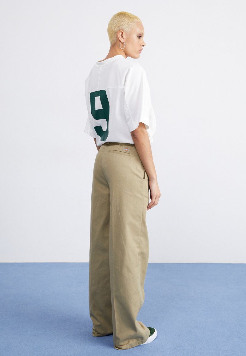 Weißes Übergröße-T-Shirt mit grüner Nummer 9 auf dem Rücken, kombiniert mit beigen weit geschnittenen Hosen. Hat eine lässige Silhouette und Taschen.